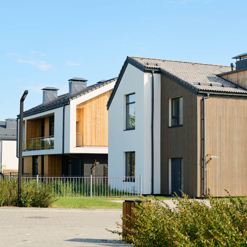 Two small houses with front yards with fencing and green bushes standing next to one another against clear blue sky